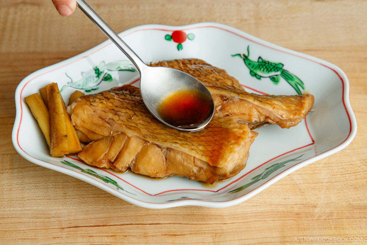 A spoon pours sauce over two pieces of cooked fish on a decorative plate, with two pieces of simmered root vegetable on the side. The plate features colorful fish illustrations and rests on a wooden surface.