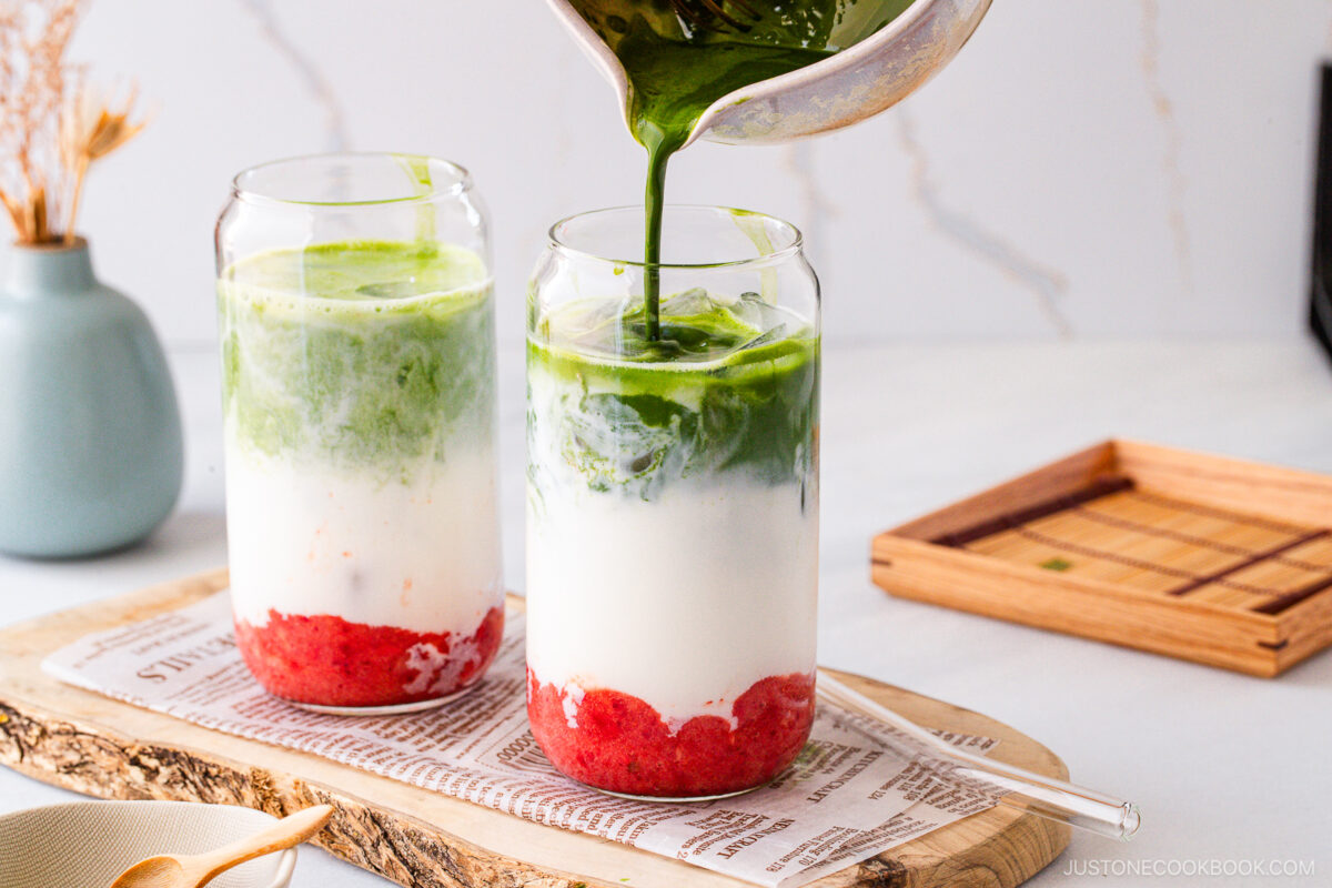 Two tall glasses filled with layers of crushed strawberries, milk, and ice are shown as a hand pours a stream of vibrant green matcha into one glass. The drinks sit on a wooden tray with a decorative background.