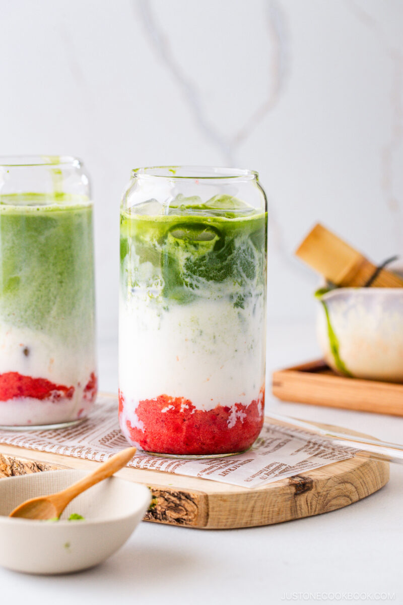 A glass filled with layers of red fruit puree, white milk, and green matcha sits on a wooden tray. Another similar glass and matcha preparation tools are in the background on a light-colored surface.