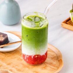 A glass of iced matcha latte with green, white, and red layers, served with a clear straw on a wooden tray. Next to it is a small dessert on a plate with a wooden pick. A matcha whisk and bowl are in the background.