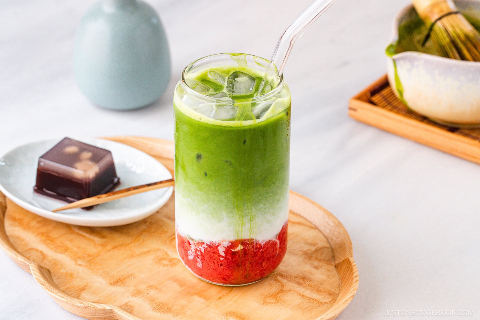 A glass of iced matcha latte with green, white, and red layers, served with a clear straw on a wooden tray. Next to it is a small dessert on a plate with a wooden pick. A matcha whisk and bowl are in the background.