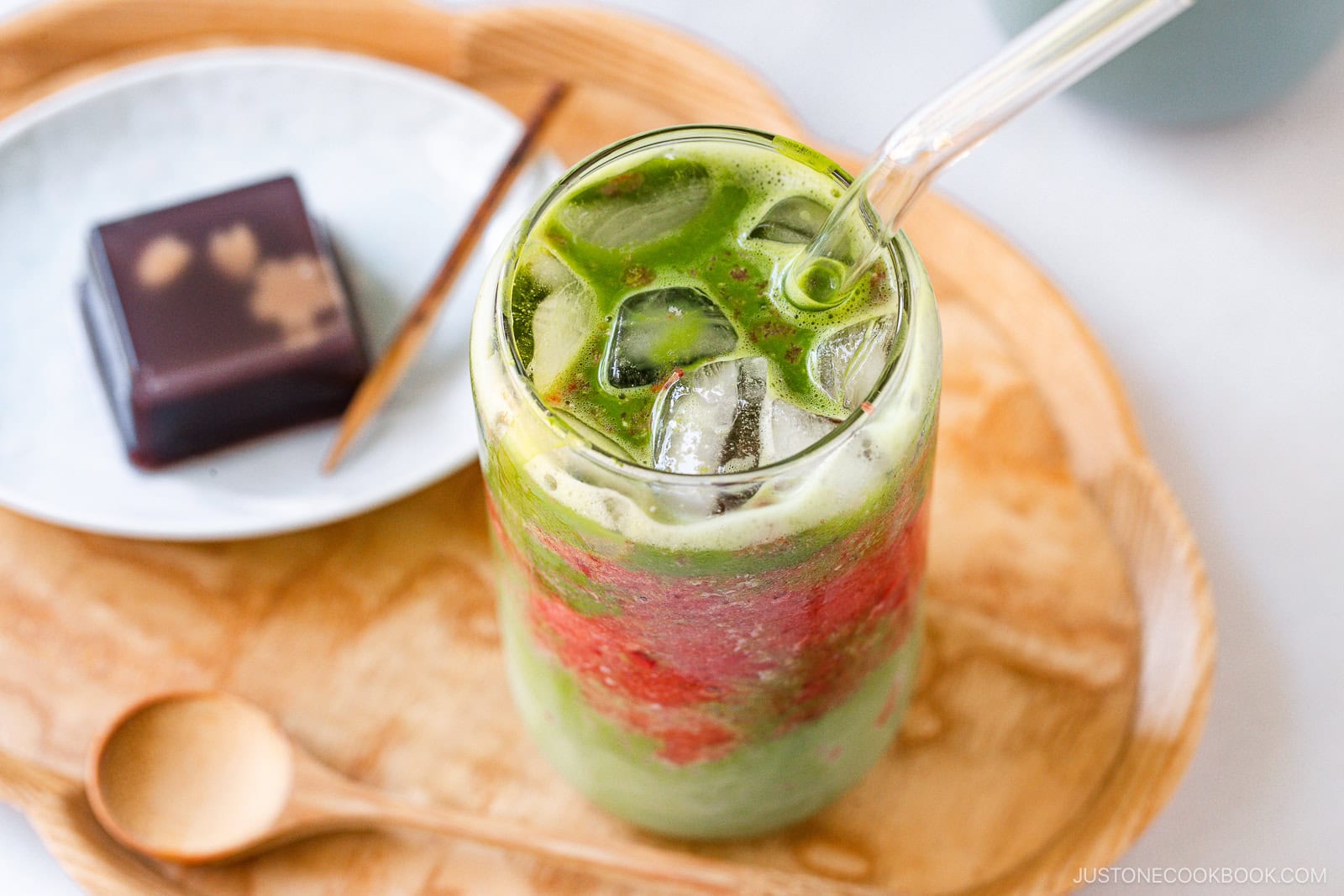 A glass of iced matcha latte with swirls of red bean and milk, served with a clear straw on a wooden tray next to a dessert and a wooden spoon.