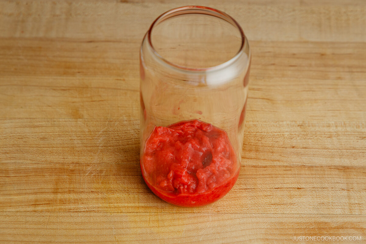 An empty glass jar with a small amount of crushed tomatoes at the bottom, sitting on a wooden surface.