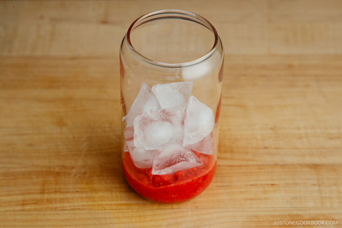 A clear glass jar with ice cubes and a layer of red fruit puree at the bottom, placed on a wooden surface.