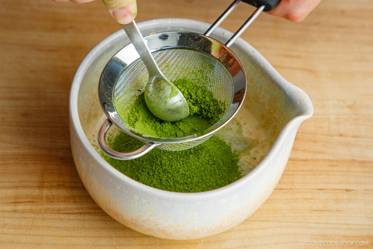 A hand holds a spoon over a fine mesh strainer with vibrant green matcha powder, sifting it into a light-colored mixing bowl on a wooden surface.