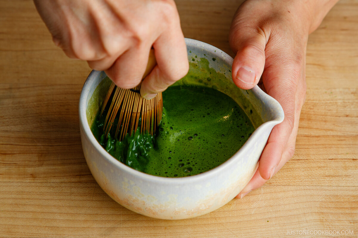A person uses a bamboo whisk to mix frothy green matcha tea in a ceramic bowl on a wooden surface.