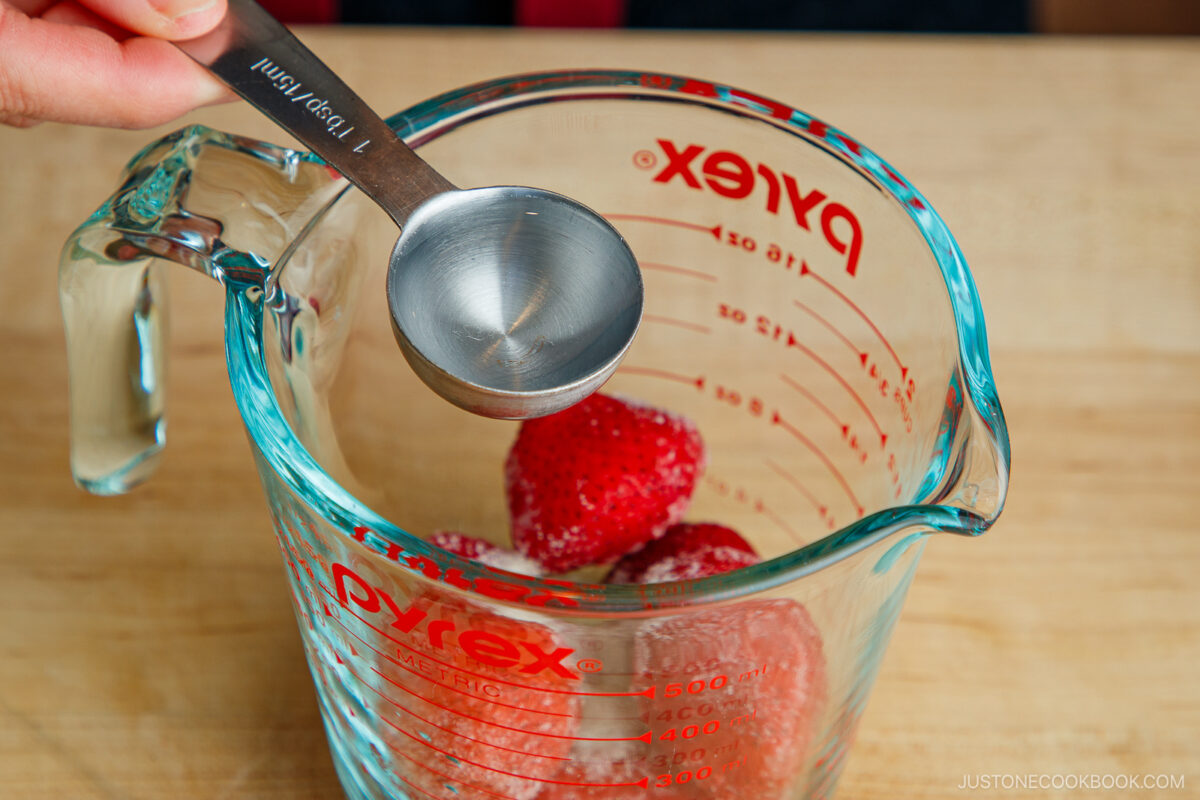 A hand holds a metal tablespoon over a glass Pyrex measuring cup containing several frozen strawberries, on a wooden surface.