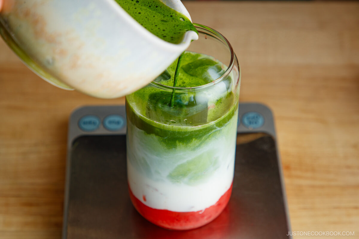 A hand pours vibrant green matcha over a glass with layered red, white, and green drinks, creating a colorful iced matcha latte on a digital kitchen scale.