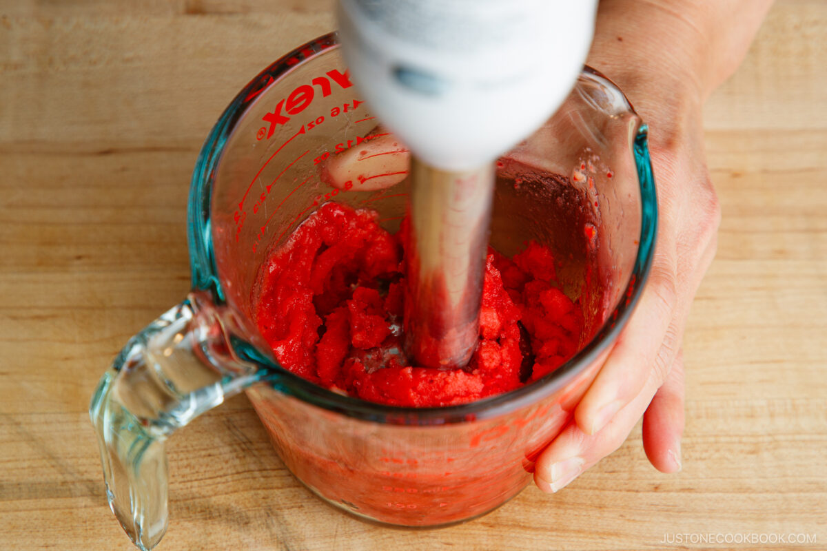 A hand holds a glass measuring cup filled with a red frozen mixture while blending it with an immersion blender on a wooden surface.