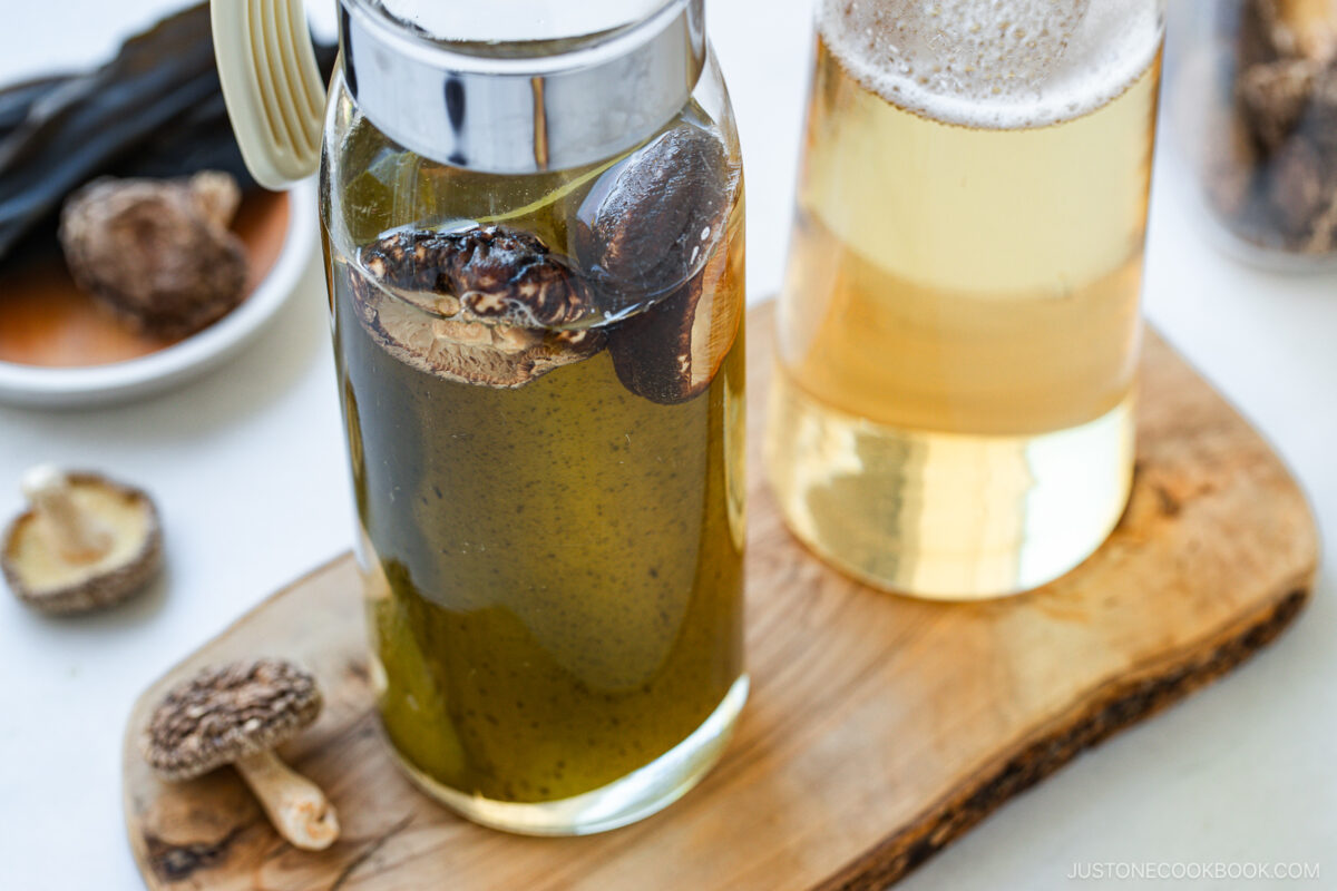 Two glass pitchers on a wooden board, one filled with clear broth and the other with a greenish broth containing dried shiitake mushrooms. More dried mushrooms are on a small plate and the board nearby.