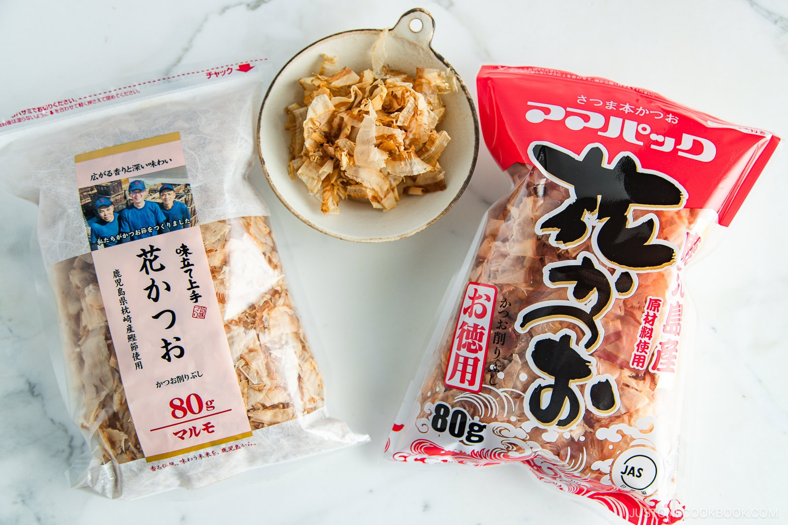Three packages of katsuobushi (dried bonito flakes) are displayed on a white surface, with two bags of flakes and a small bowl containing some of the flakes in the center. The packaging features Japanese text.