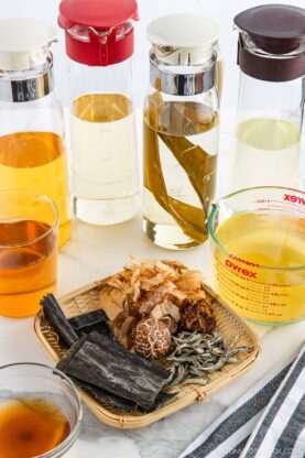 A selection of glass pitchers with light-colored broths, a measuring cup with liquid, and a bamboo tray displaying dried seaweed, mushrooms, bonito flakes, and other dashi ingredients on a white surface.