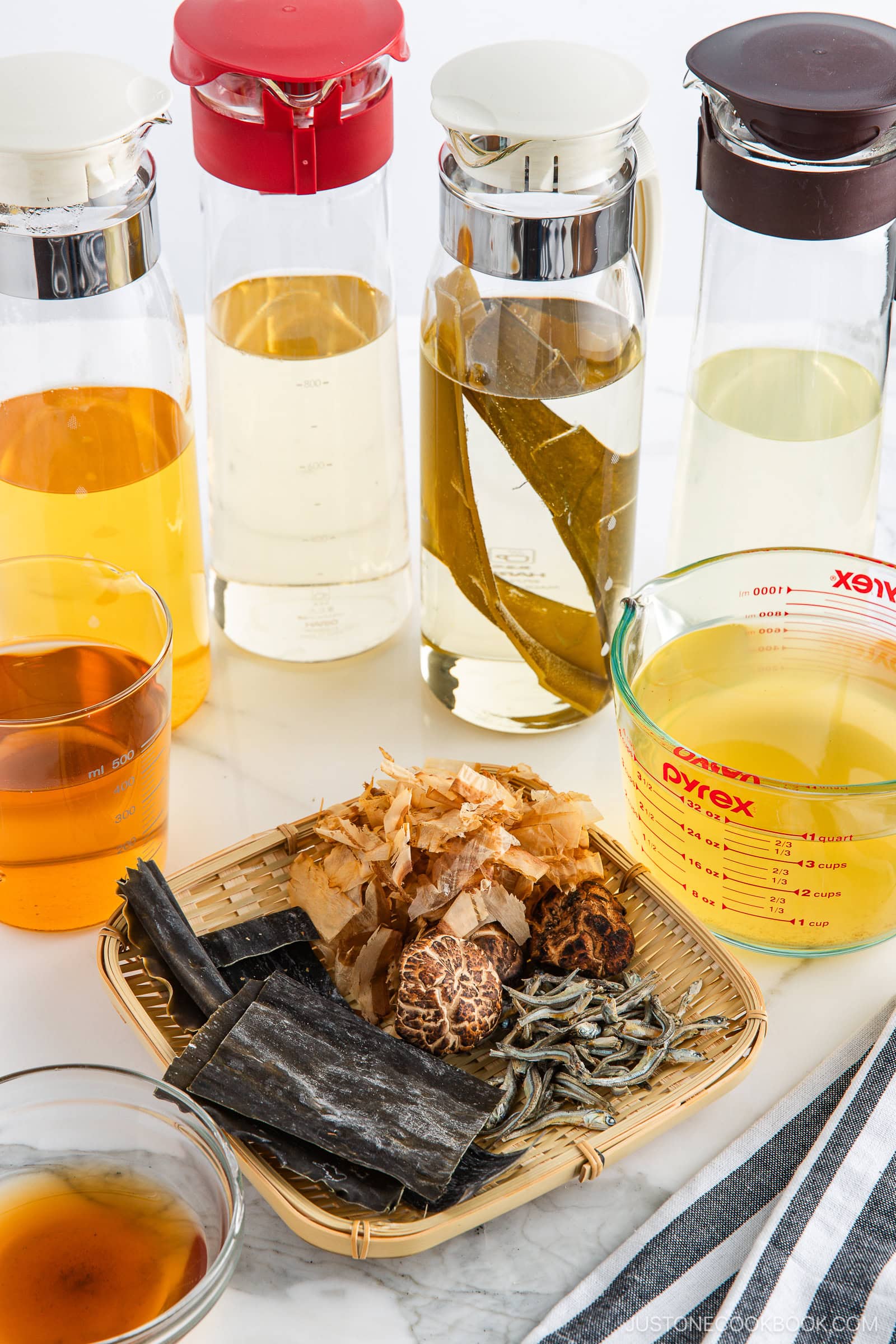 A selection of glass pitchers with light-colored broths, a measuring cup with liquid, and a bamboo tray displaying dried seaweed, mushrooms, bonito flakes, and other dashi ingredients on a white surface.