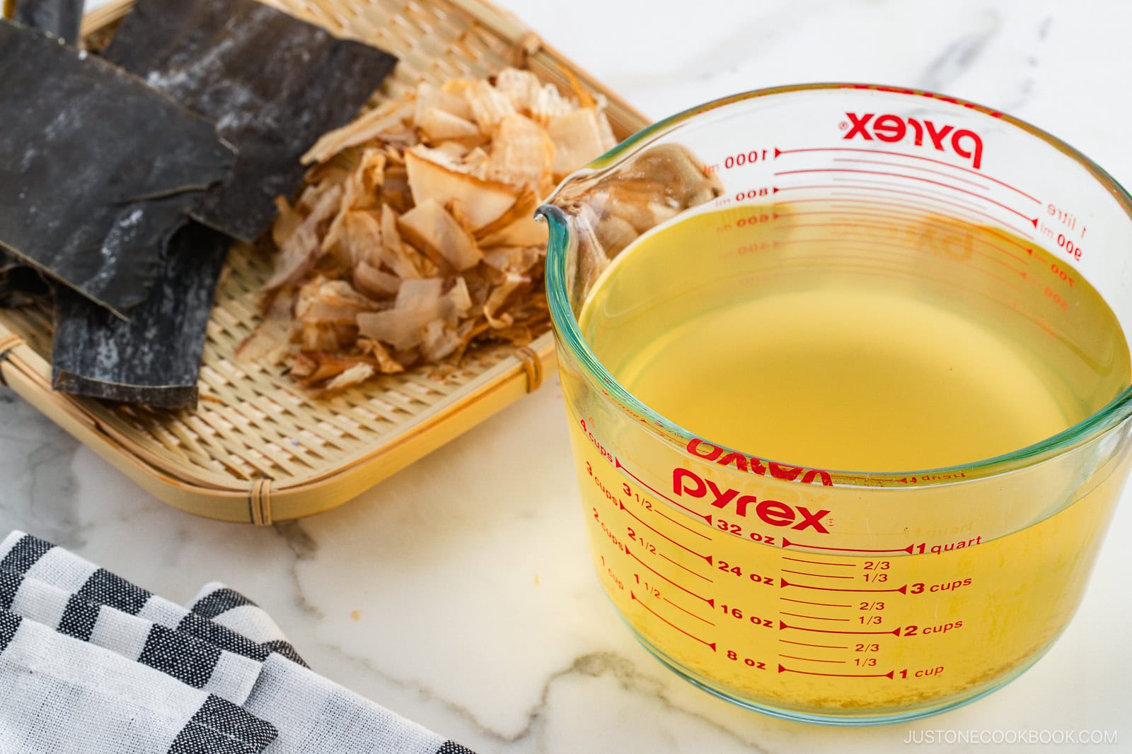 A glass measuring cup filled with clear golden dashi broth sits on a white surface. Nearby, a bamboo tray holds dry kombu seaweed and bonito flakes. A black and white cloth is partially visible in the corner.