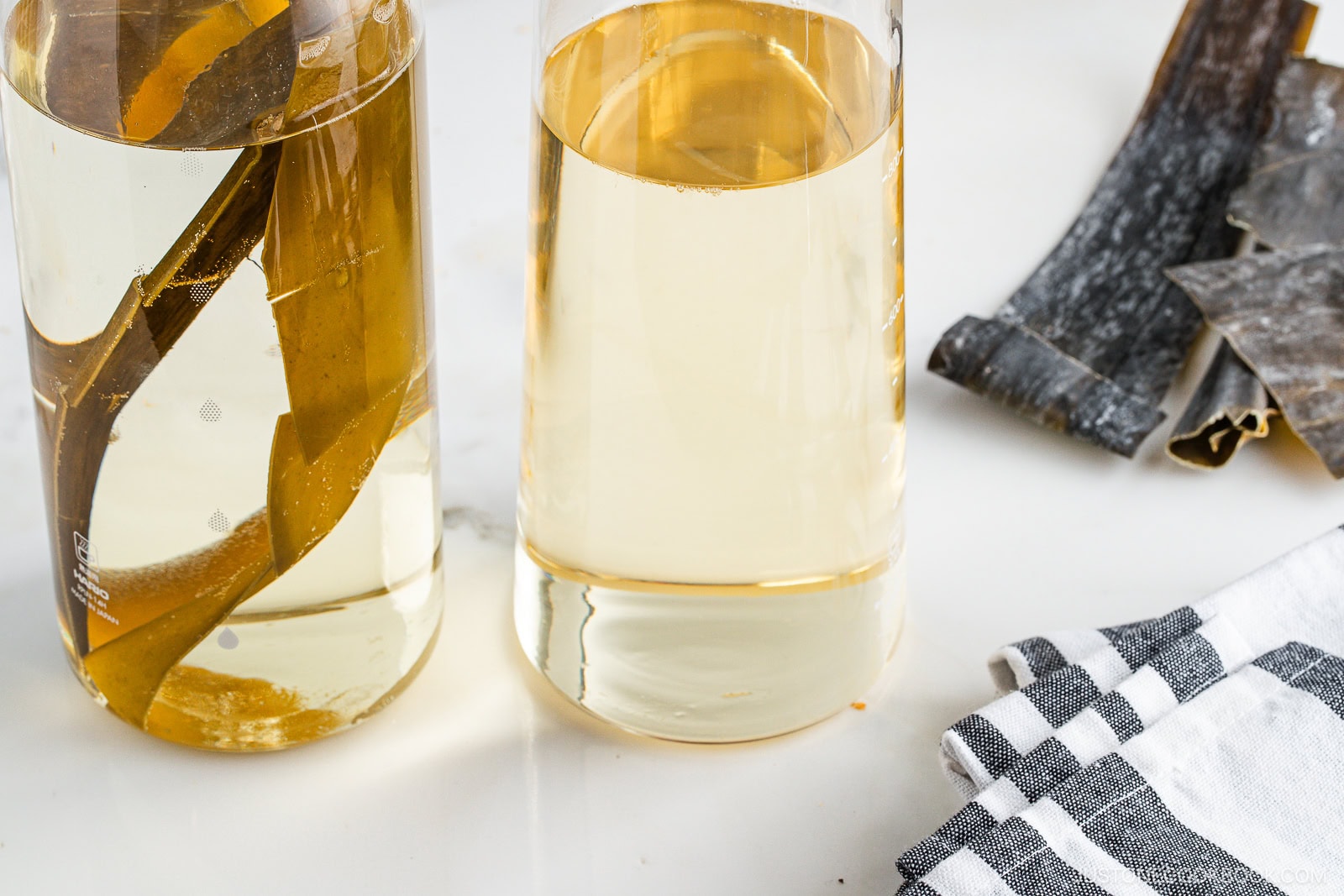 Two glass containers filled with light golden liquid and pieces of kombu (seaweed) inside, placed on a white surface. Dried kombu sheets and a folded black-and-white checkered cloth are nearby.