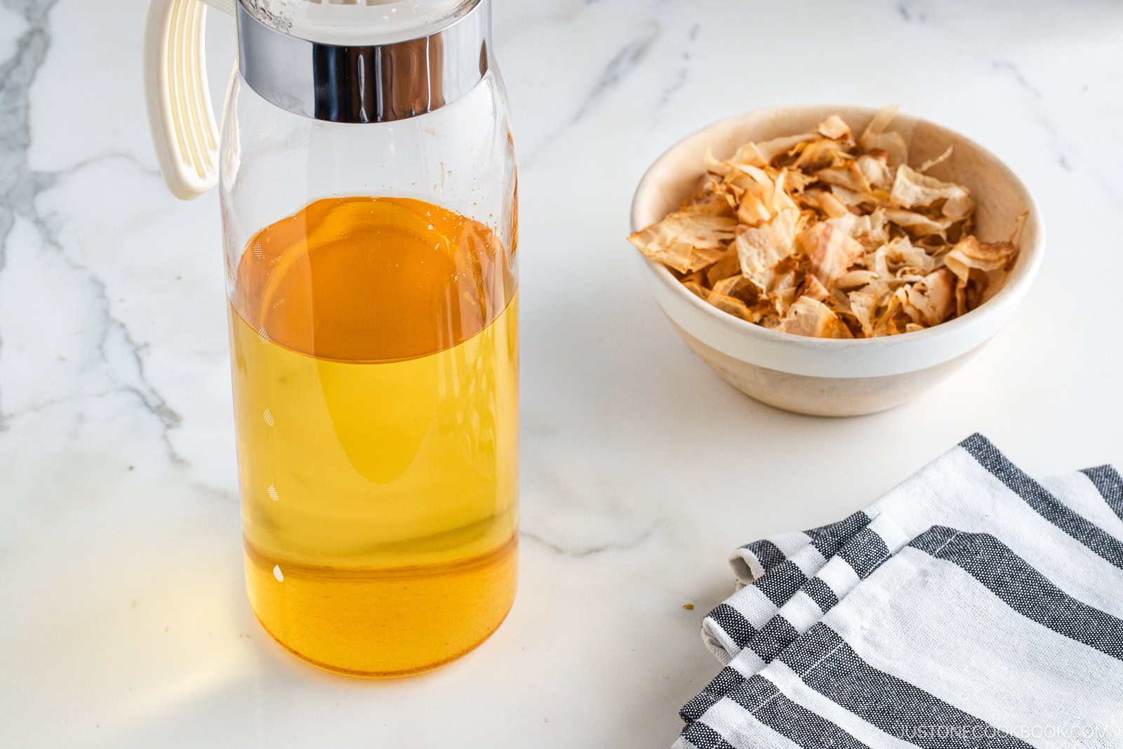A glass pitcher filled with golden liquid stands next to a bowl of dried onion skins on a white marble surface with a black-and-white striped cloth nearby.