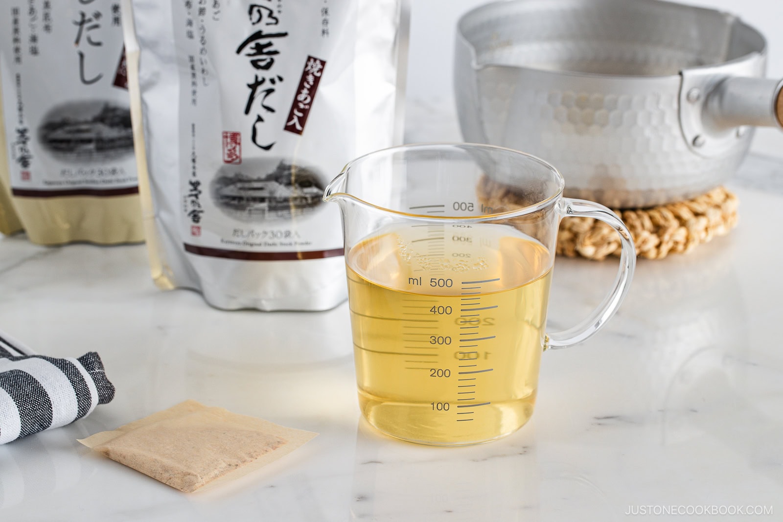 A clear measuring cup filled with light golden liquid sits on a white surface, surrounded by a tea bag, packaged ingredients, a striped towel, and a metal pot on a woven trivet in the background.