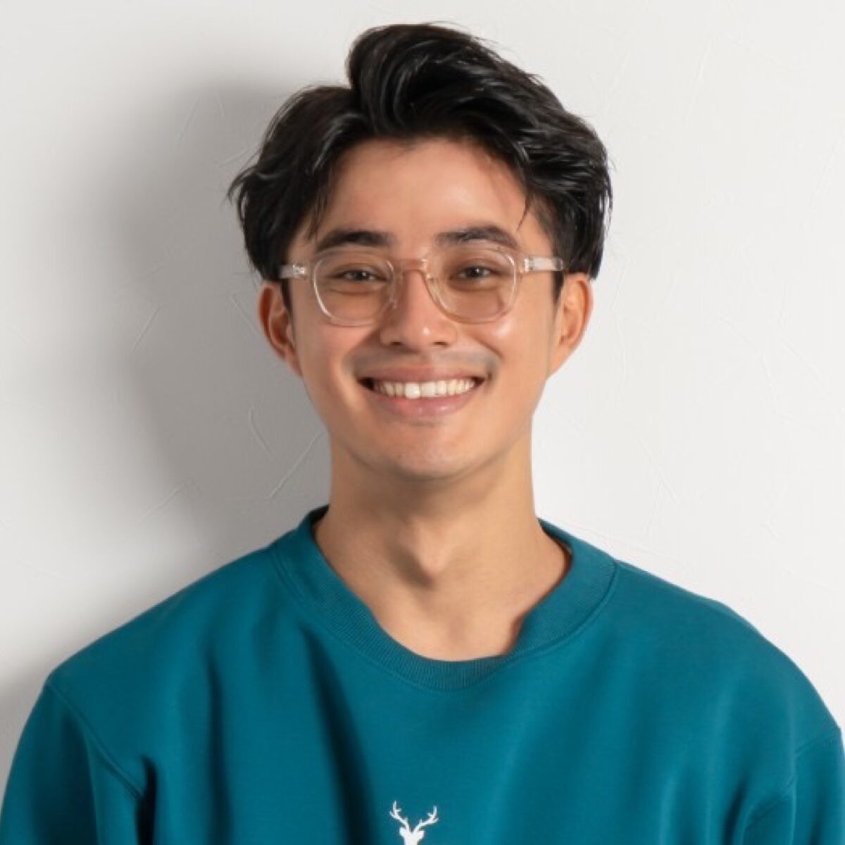 Young Asian man with black hair - smiling, wearing clear glasses and a teal sweatshirt, portrait shot in front of plain white background.