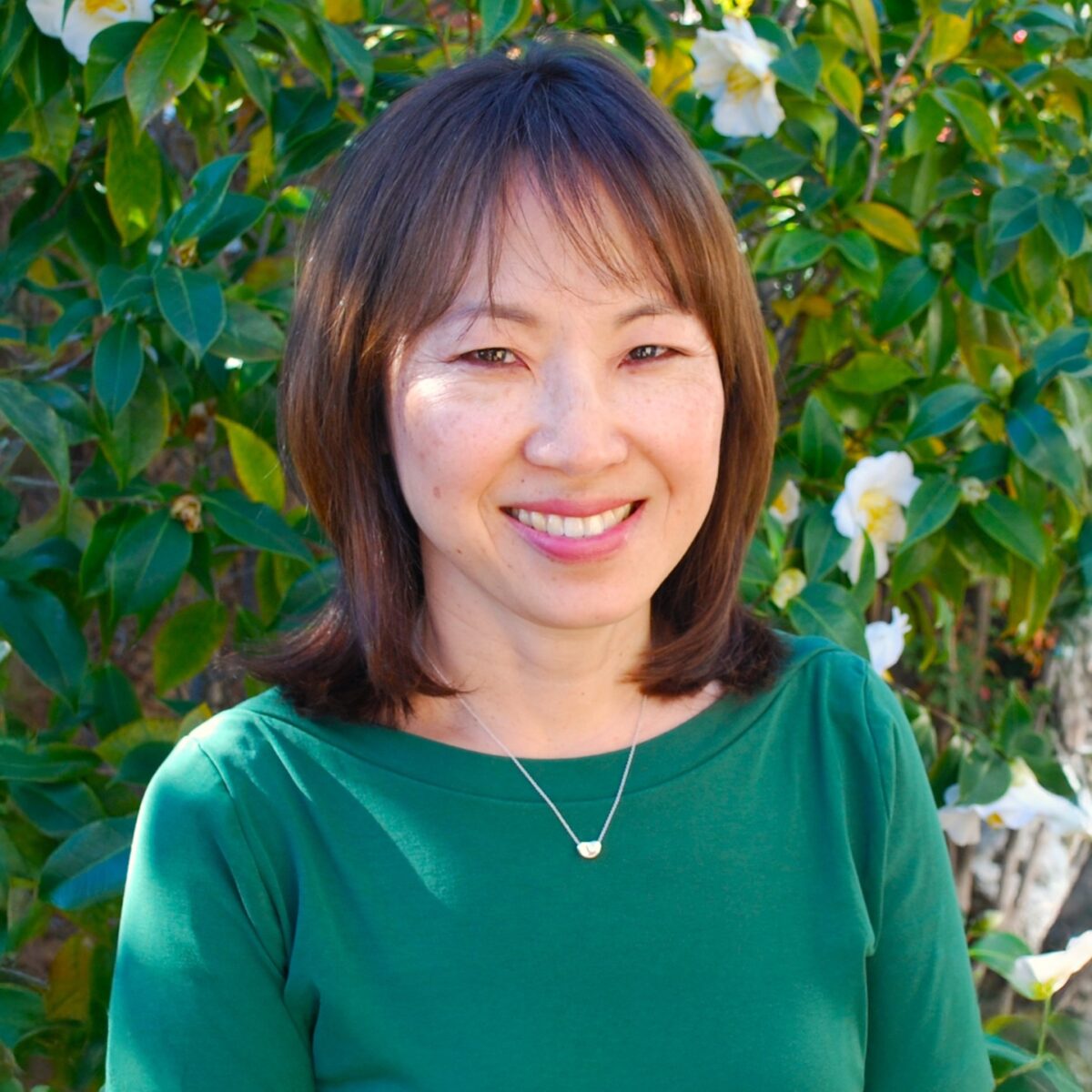 Smiling woman outdoors with lush green leaves and white flowers, showcasing a friendly personality, suitable for content on Japanese cuisine, cooking tips, or food culture.