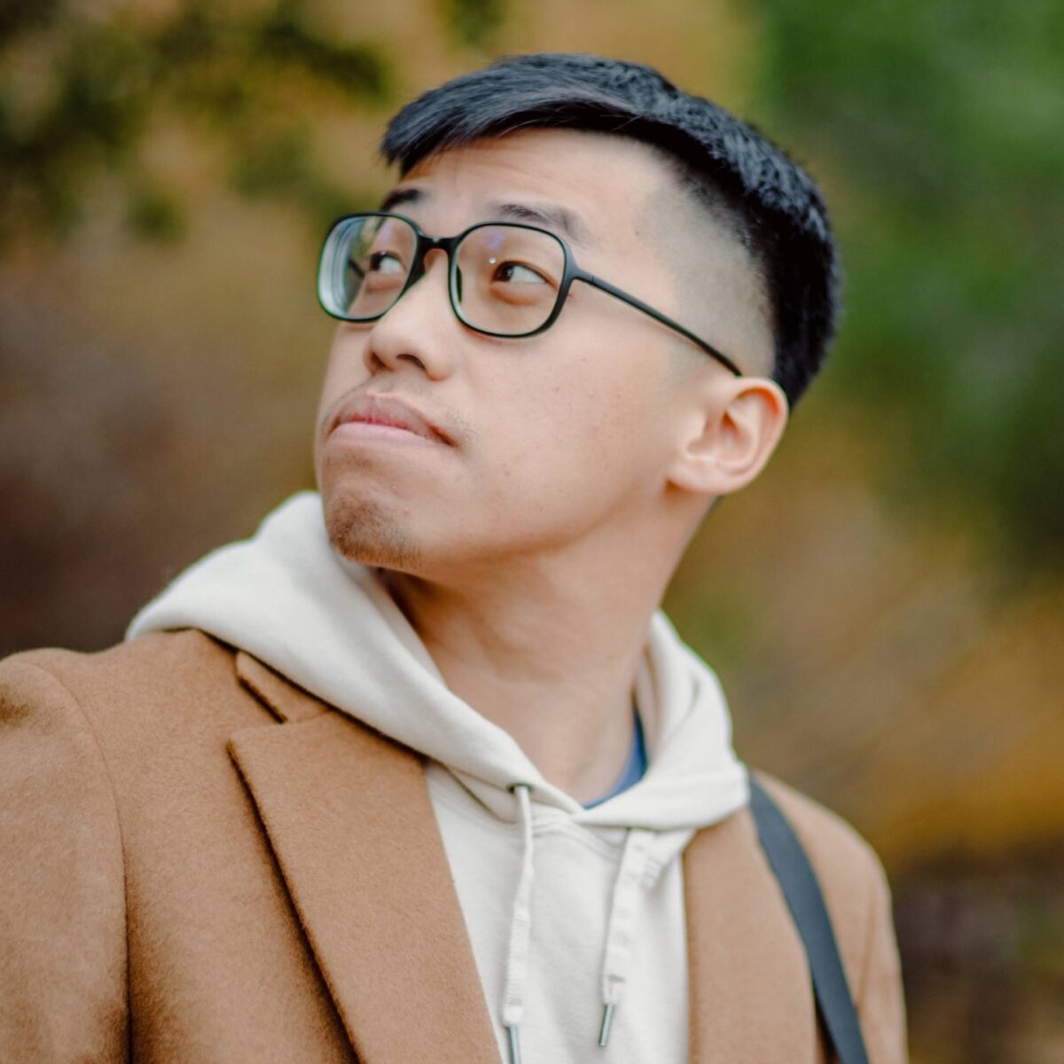 Stylish Asian man wearing glasses, beige coat, and hoodie outdoors, showcasing fashion and lifestyle.