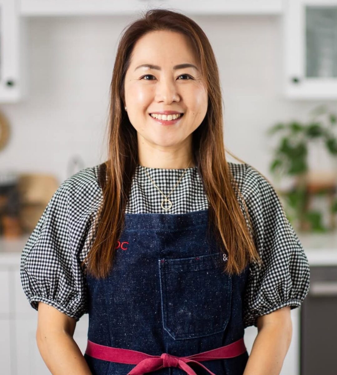 Smiling woman in a kitchen wearing a black and white checkered top and navy apron, showcasing Japanese home cooking and culinary expertise.