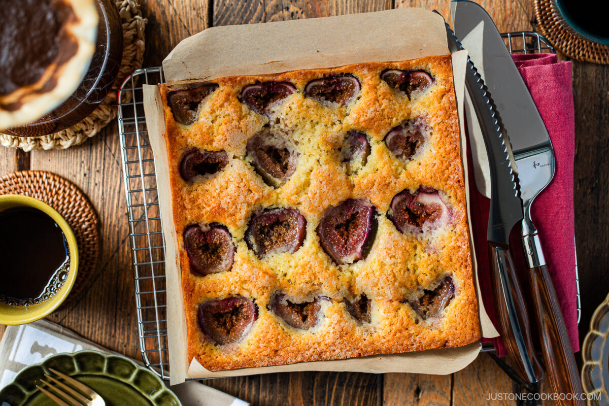 A freshly baked fig cake in a square pan sits on a cooling rack, with visible fig halves on top. Surrounding the cake are plates, utensils, a cup of coffee, and a pie, all on a rustic wooden table.
