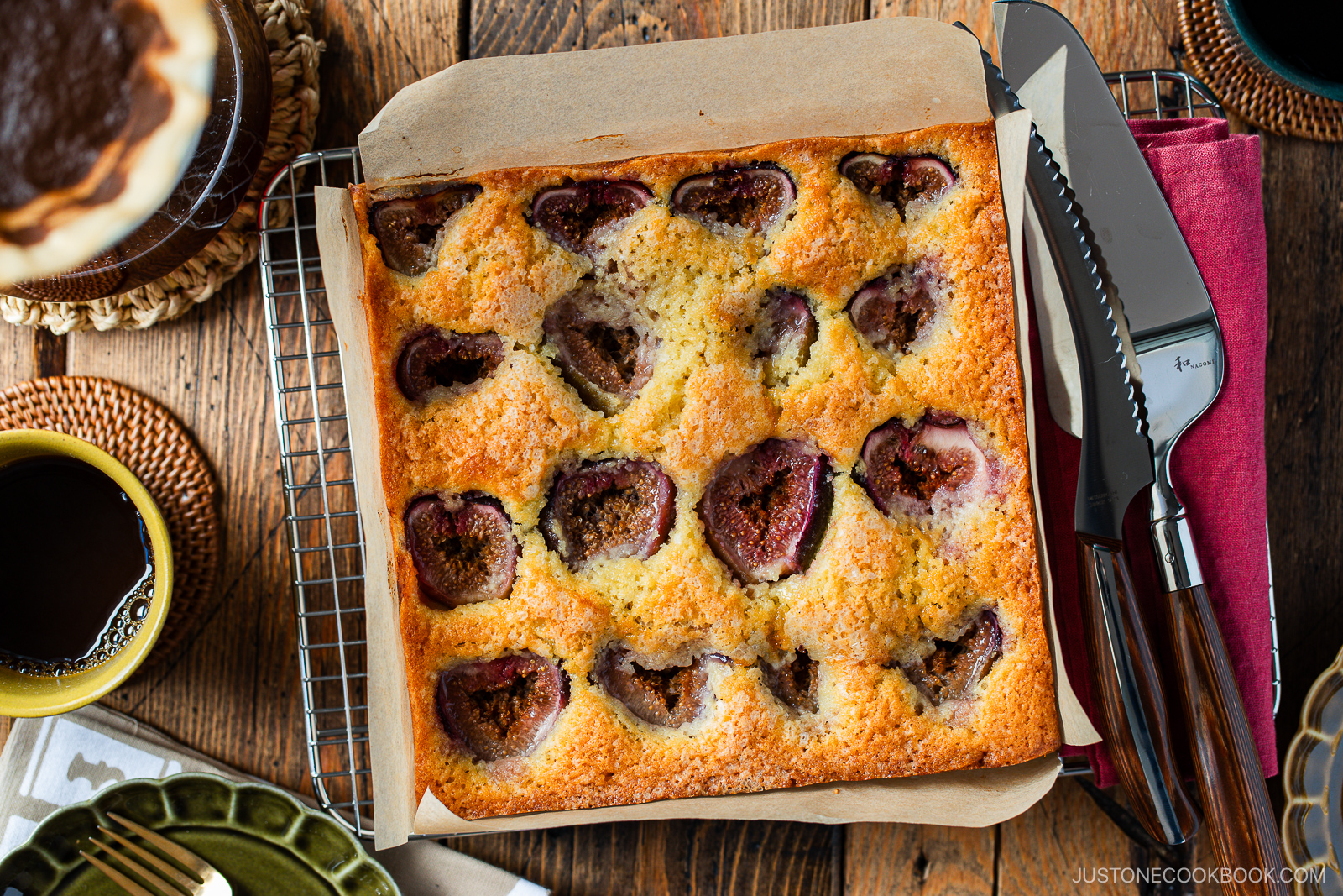A freshly baked fig cake in a square pan sits on a cooling rack, with visible fig halves on top. Surrounding the cake are plates, utensils, a cup of coffee, and a pie, all on a rustic wooden table.
