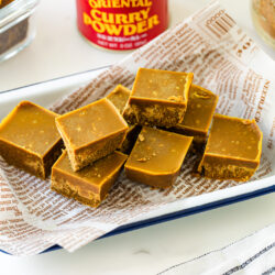 A white enamel tray lined with printed paper holds several pieces of golden brown fudge. A jar labeled Oriental Curry Powder and a glass container of more fudge are in the background.