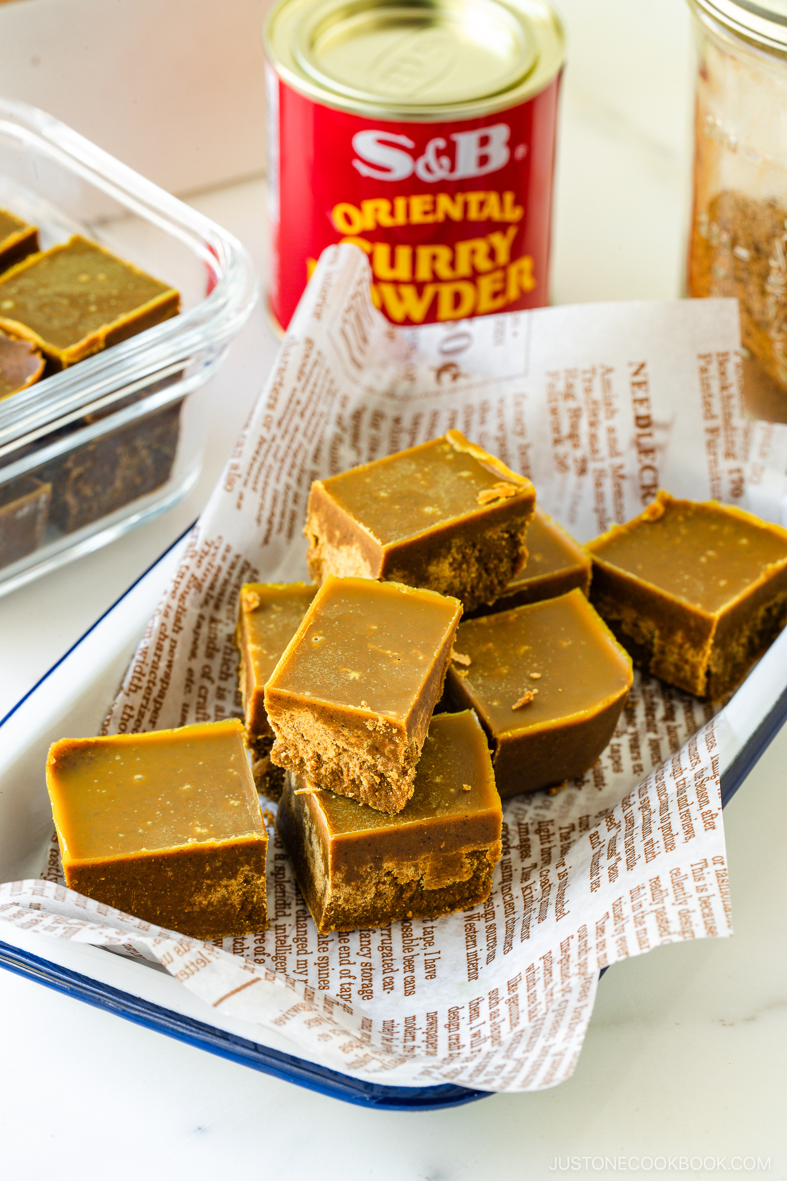 A tray lined with printed paper holds several square pieces of curry fudge. A glass dish with more fudge and a can of S&B curry powder are visible in the background.