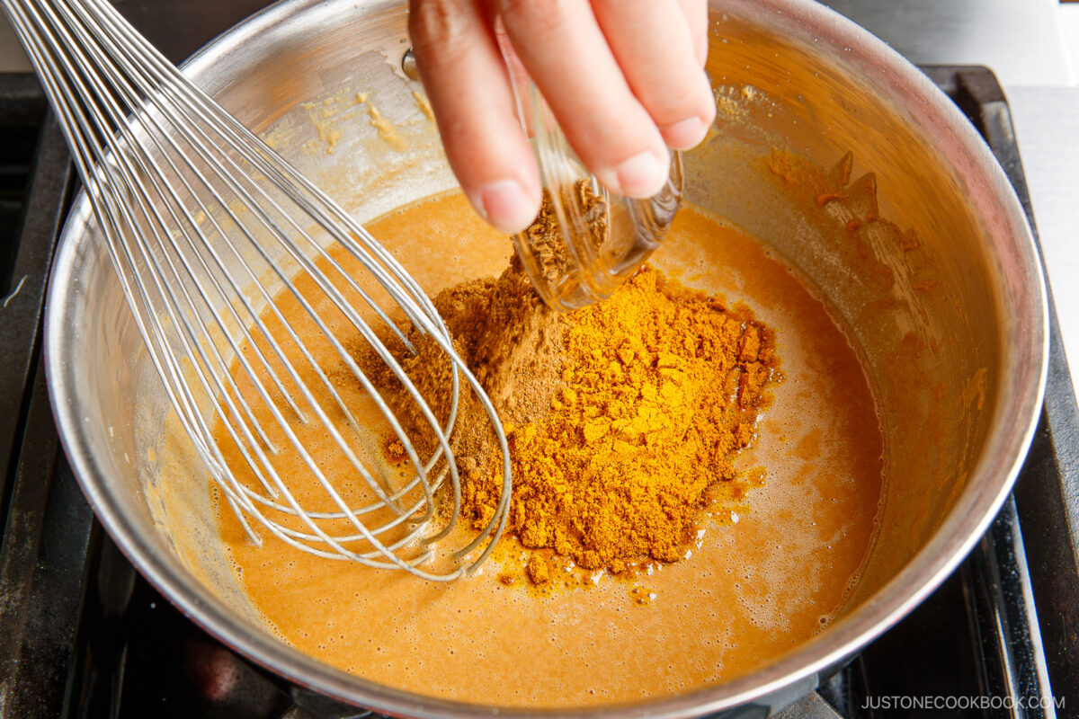A hand pours ground curry powder from a small bowl into a saucepan with a brown sauce, next to a metal whisk.