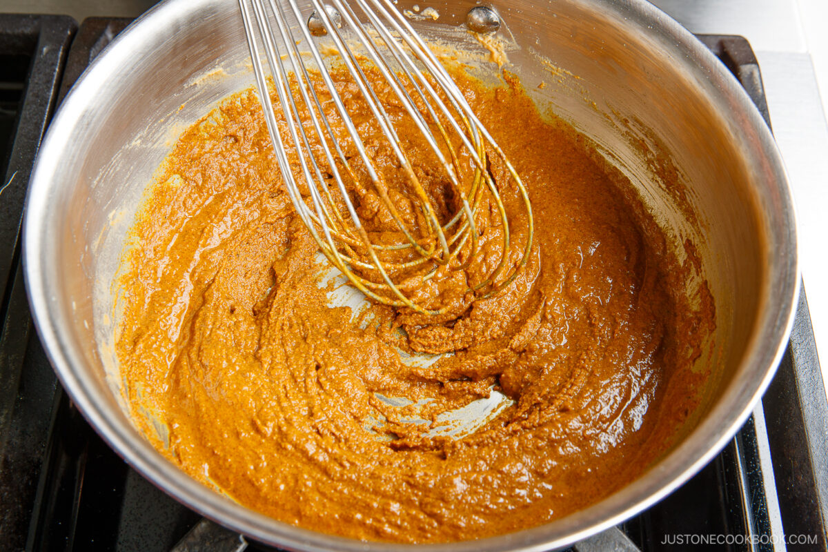 A metal whisk stirs a thick, brown curry roux in a stainless steel saucepan on a stovetop.