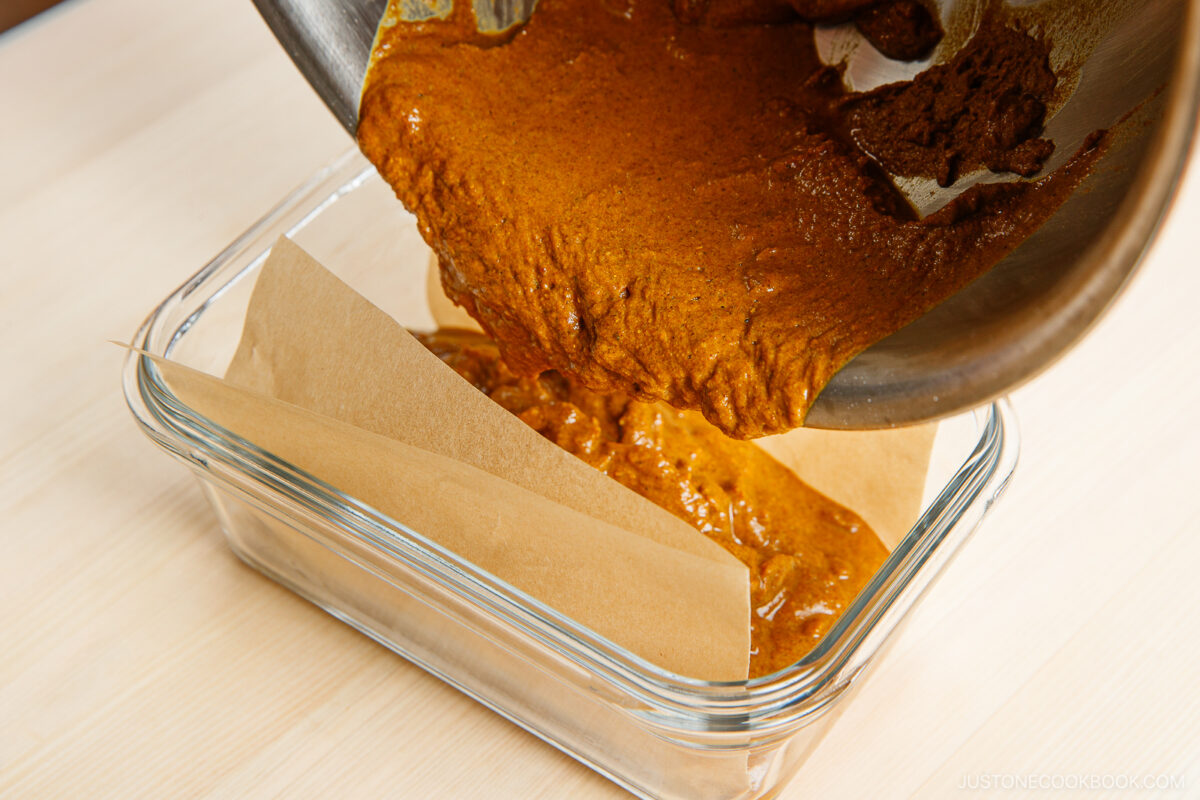 A thick brown mixture is being poured from a pan into a parchment-lined rectangular glass baking dish on a light wooden surface.