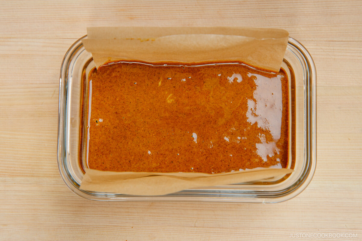 A glass dish lined with parchment paper is filled with a smooth, glossy, brown sauce or mixture, likely being prepared for baking or setting. The dish rests on a light wooden surface.
