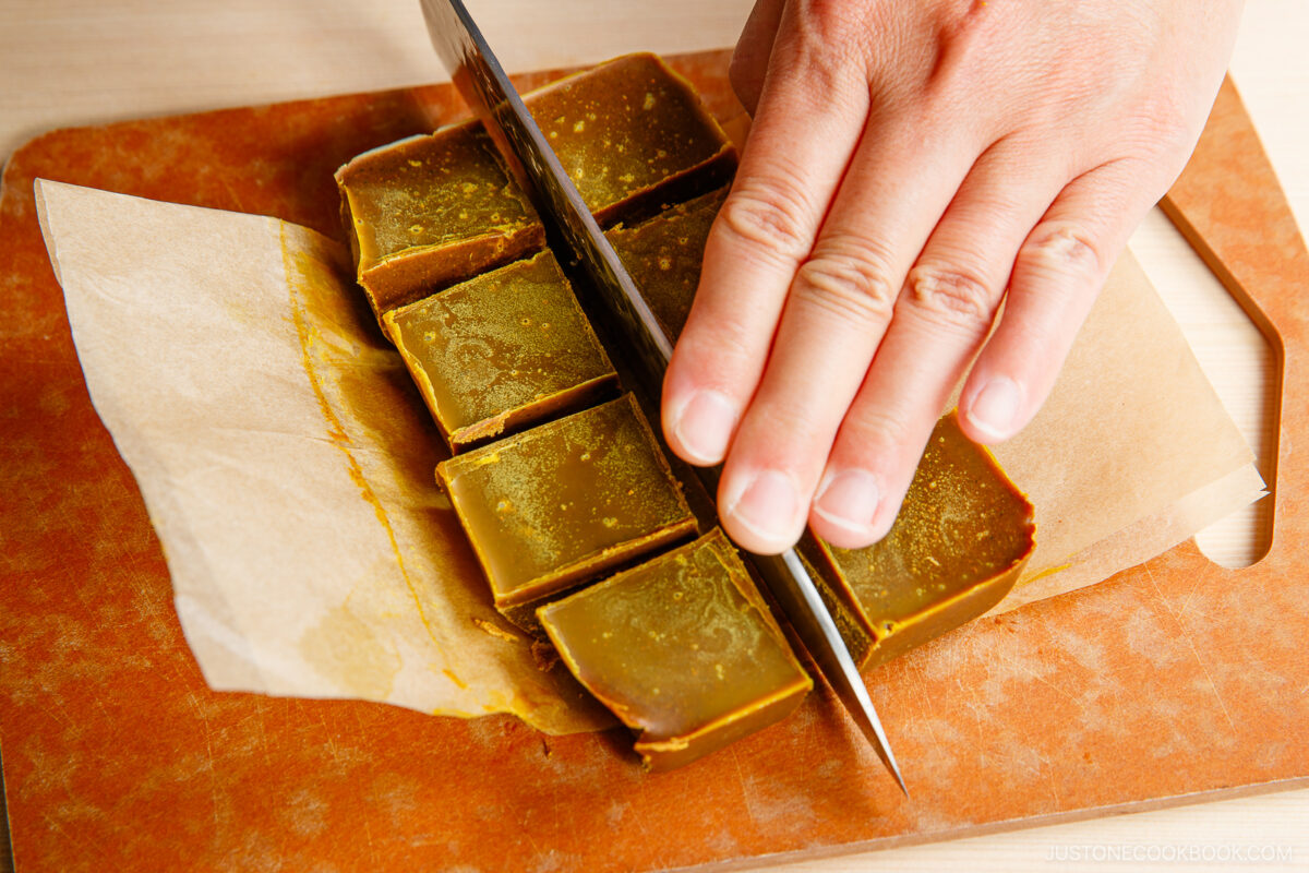 A hand slices a block of brown matcha caramel fudge into squares on a cutting board lined with parchment paper.