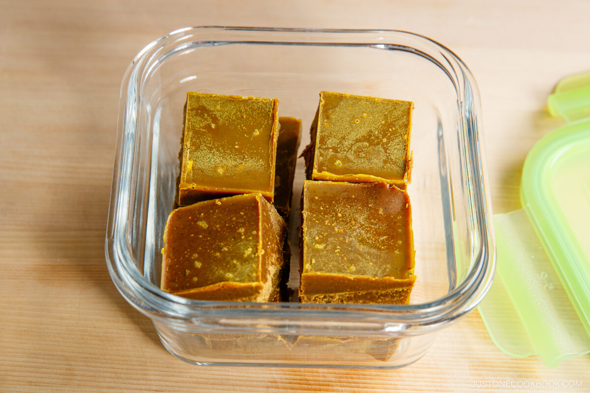 Four rectangular blocks of golden-brown Japanese curry roux are arranged in a glass container with a green lid placed beside it on a wooden surface.