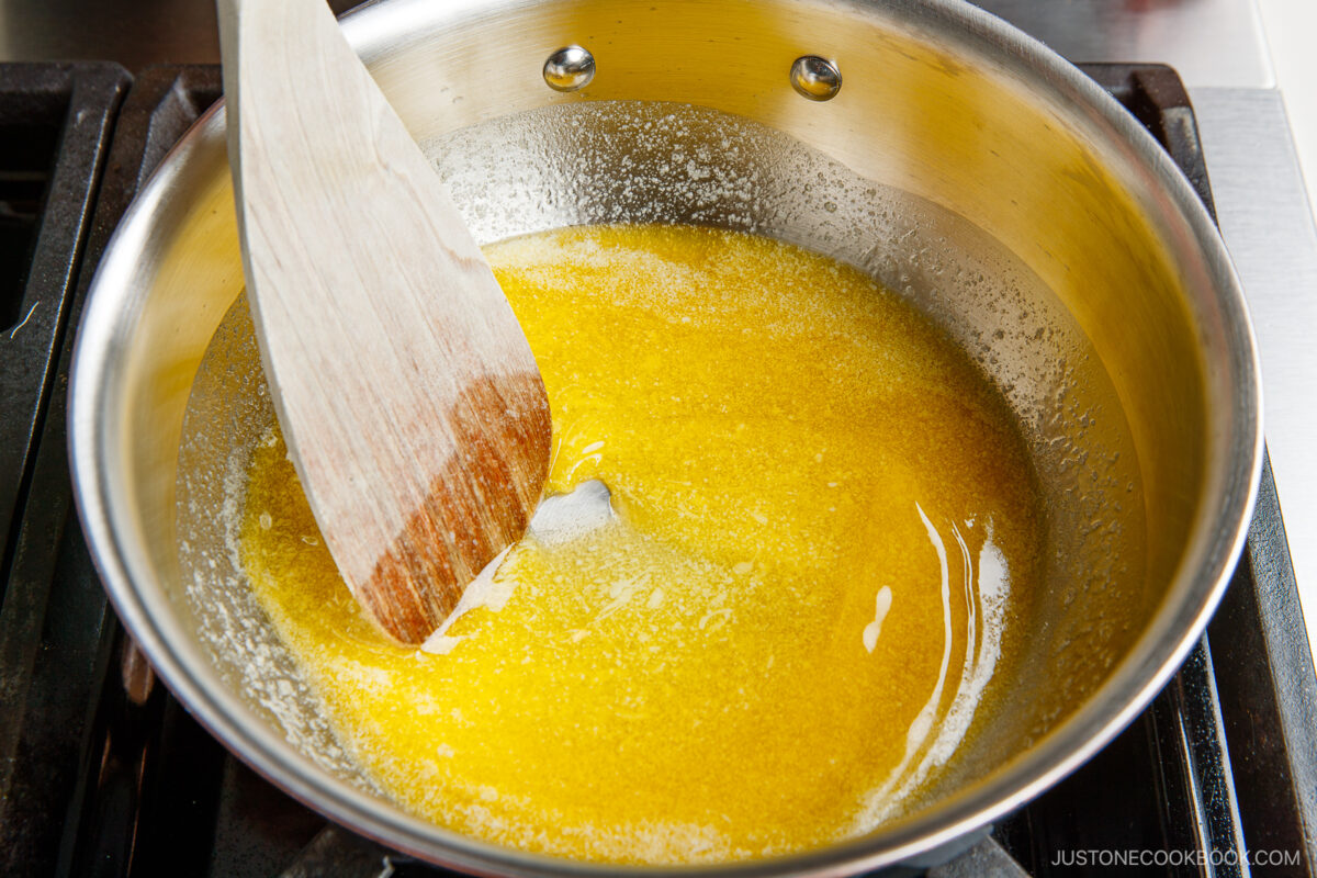 A wooden spatula stirs melted butter in a stainless steel pan on a stovetop. The butter is yellow and bubbling, indicating it is being heated or clarified.