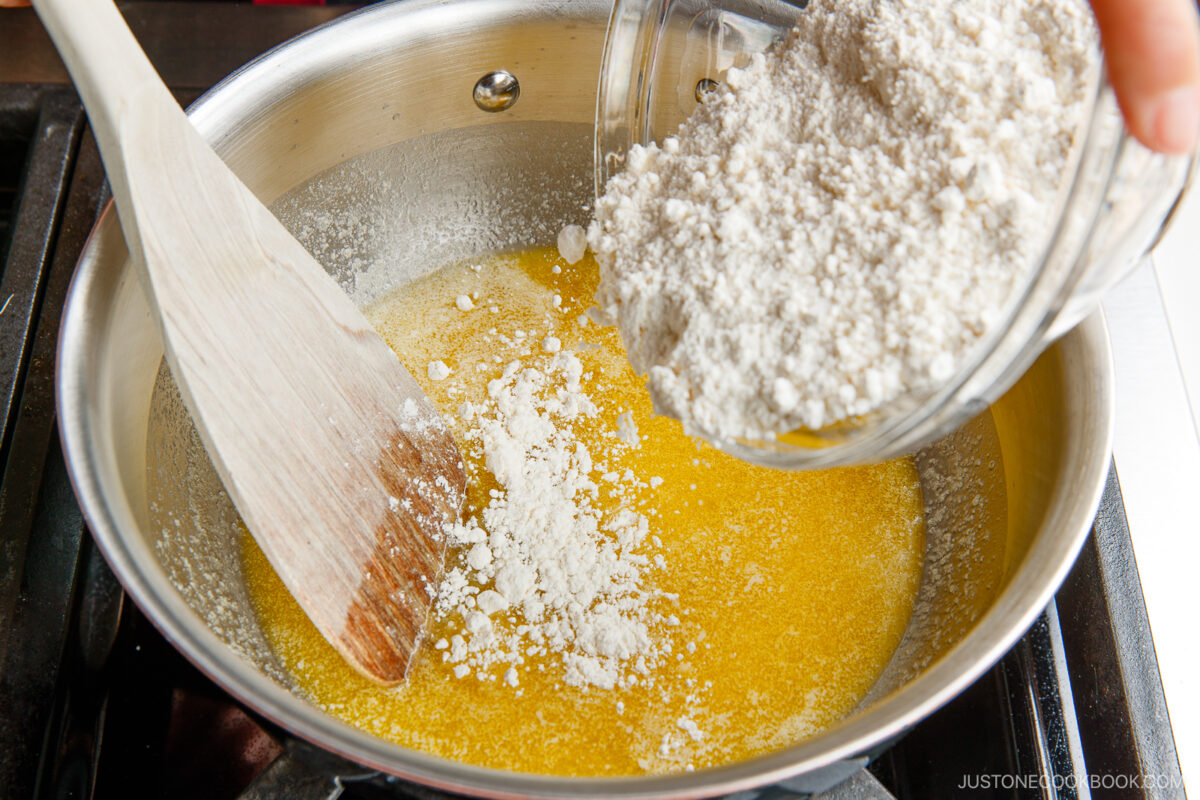 A hand pours flour from a glass container into a pan of melted butter on a stovetop, mixing with a wooden spatula.