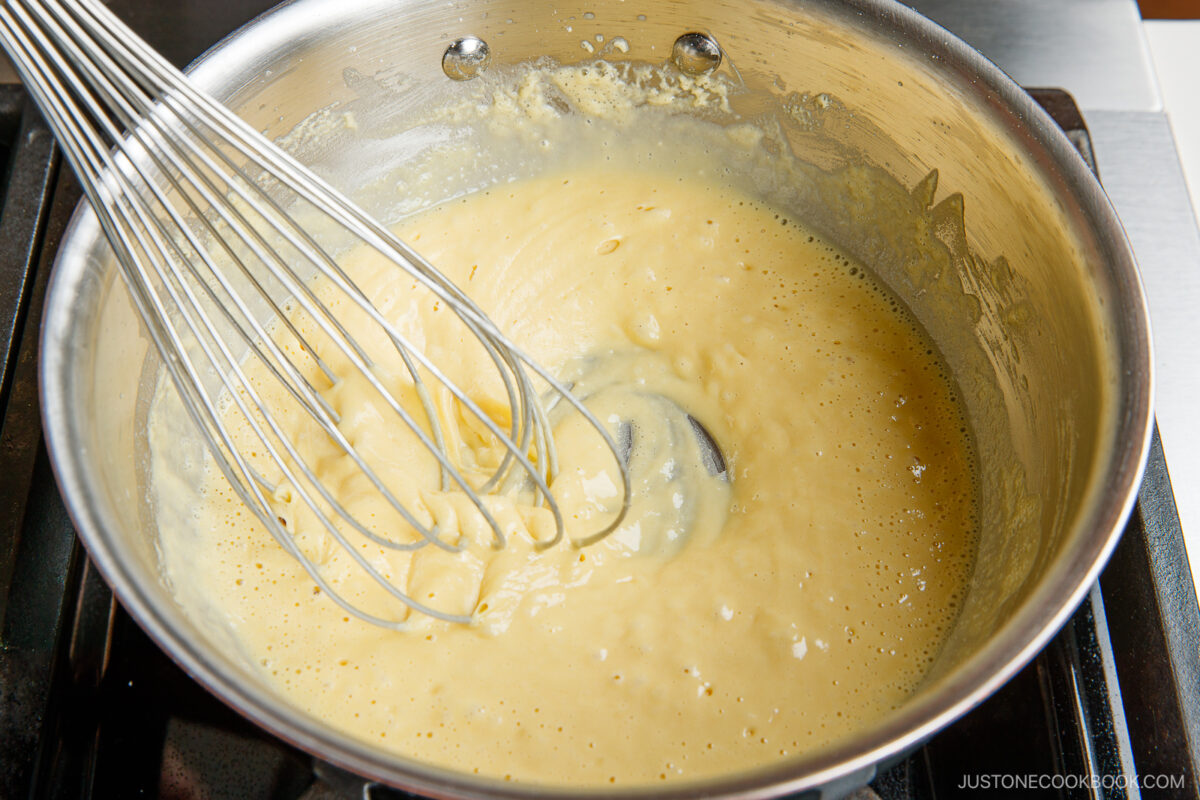 A stainless steel whisk stirring a thick, creamy sauce in a metal saucepan on a stovetop, with light bubbles forming on the surface.