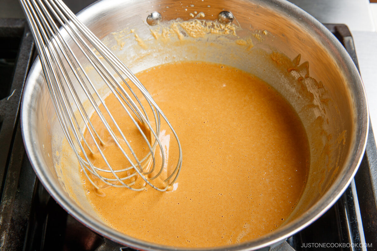 A stainless steel saucepan filled with a smooth, light brown sauce being whisked, sitting on a stovetop.