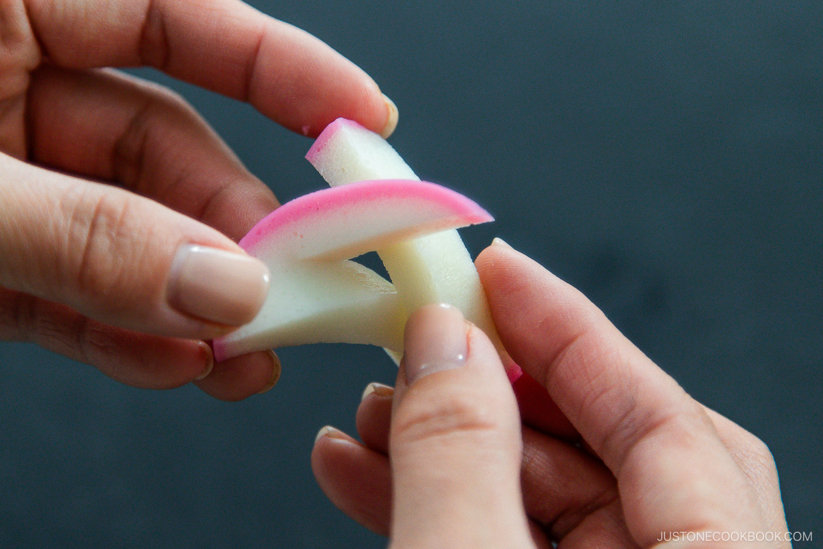 Two hands are twisting a piece of decorative pink and white pickled radish into a simple knot against a dark background.