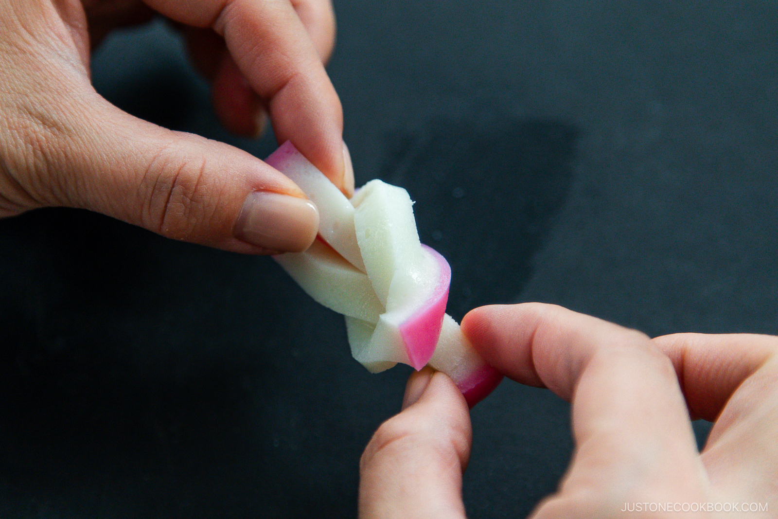 Close-up of hands tying a decorative knot with a strip of pink and white fish cake (kamaboko) on a dark surface.