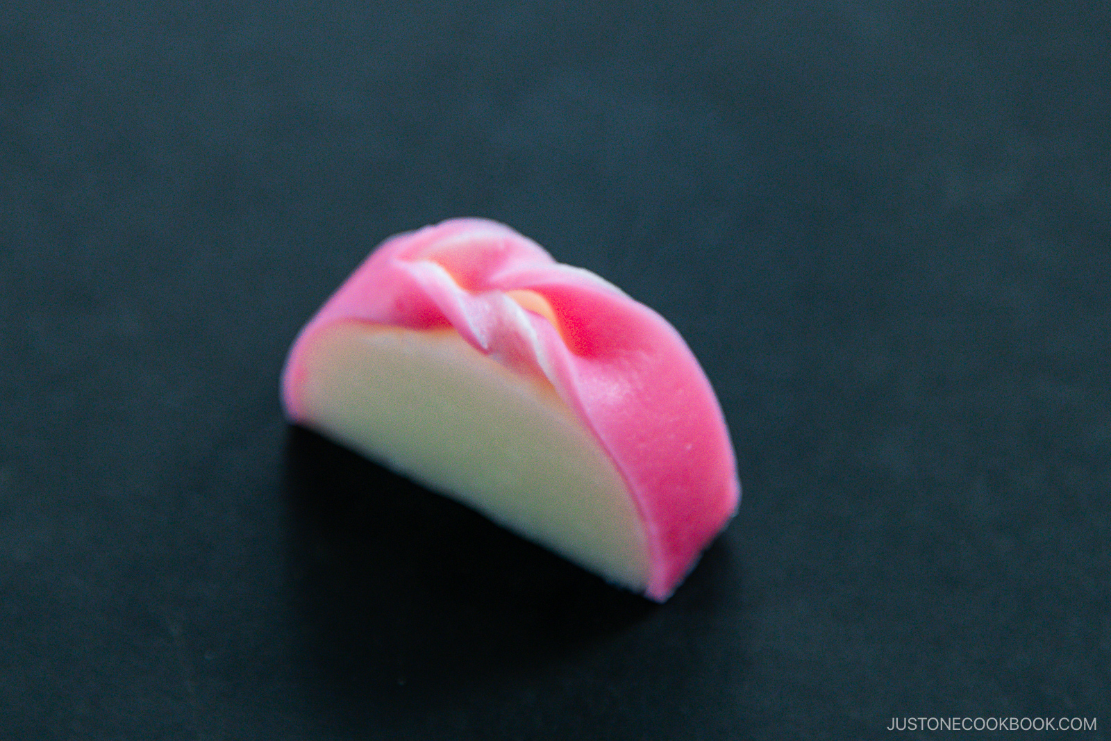 A piece of pink and white Japanese wagashi (traditional sweet) shaped like a half-moon, placed on a dark surface. The sweet has a soft, smooth texture with delicate folding at the top.
