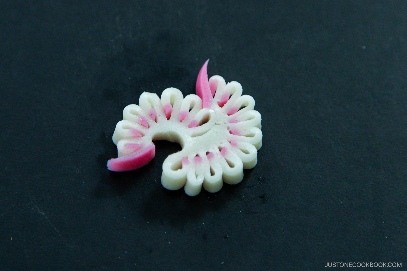 A decorative slice of Japanese fish cake (kamaboko), intricately cut with a white base and pink accents, displayed on a dark background.
