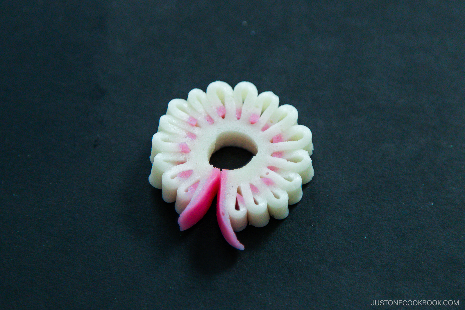 A decorative white and pink flower-shaped piece of fish cake (kamaboko) with a hole in the center, placed on a dark background.