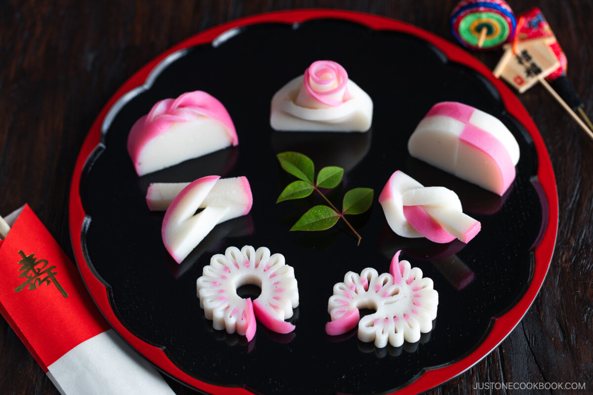 A black tray with six pieces of decorative pink and white Japanese kamaboko fish cake, arranged artistically with green leaves in the center, and chopsticks with a red paper sleeve on the side.
