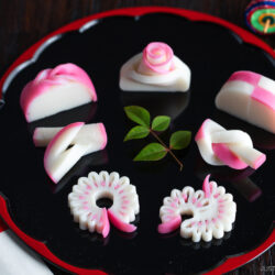 A black tray with six pieces of decorative pink and white Japanese kamaboko fish cake, arranged artistically with green leaves in the center, and chopsticks with a red paper sleeve on the side.