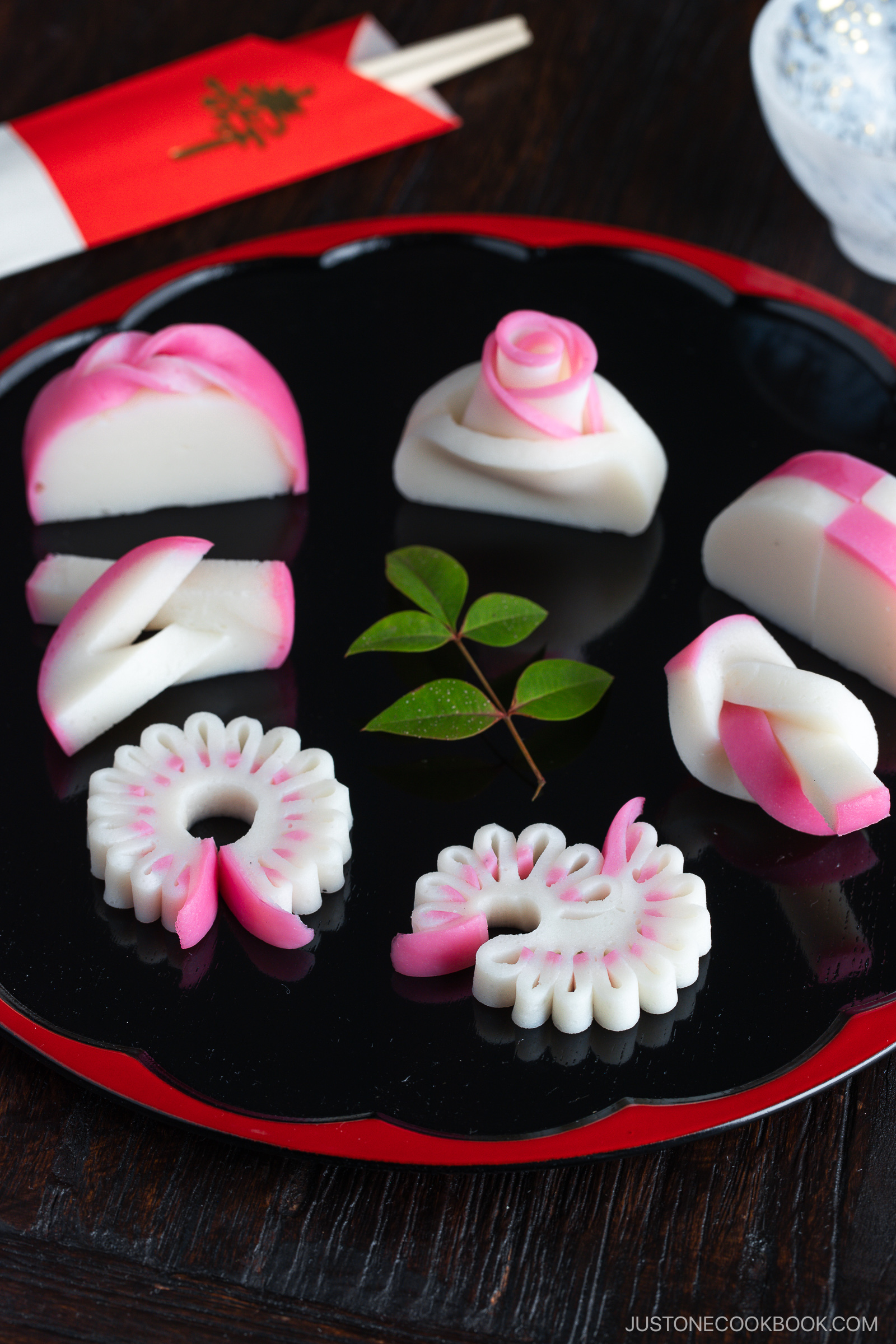 Assorted decorative slices of pink and white Japanese kamaboko (fish cake) are arranged on a black lacquer tray with a small green leaf, beside red chopsticks and a folded napkin on a dark wooden table.
