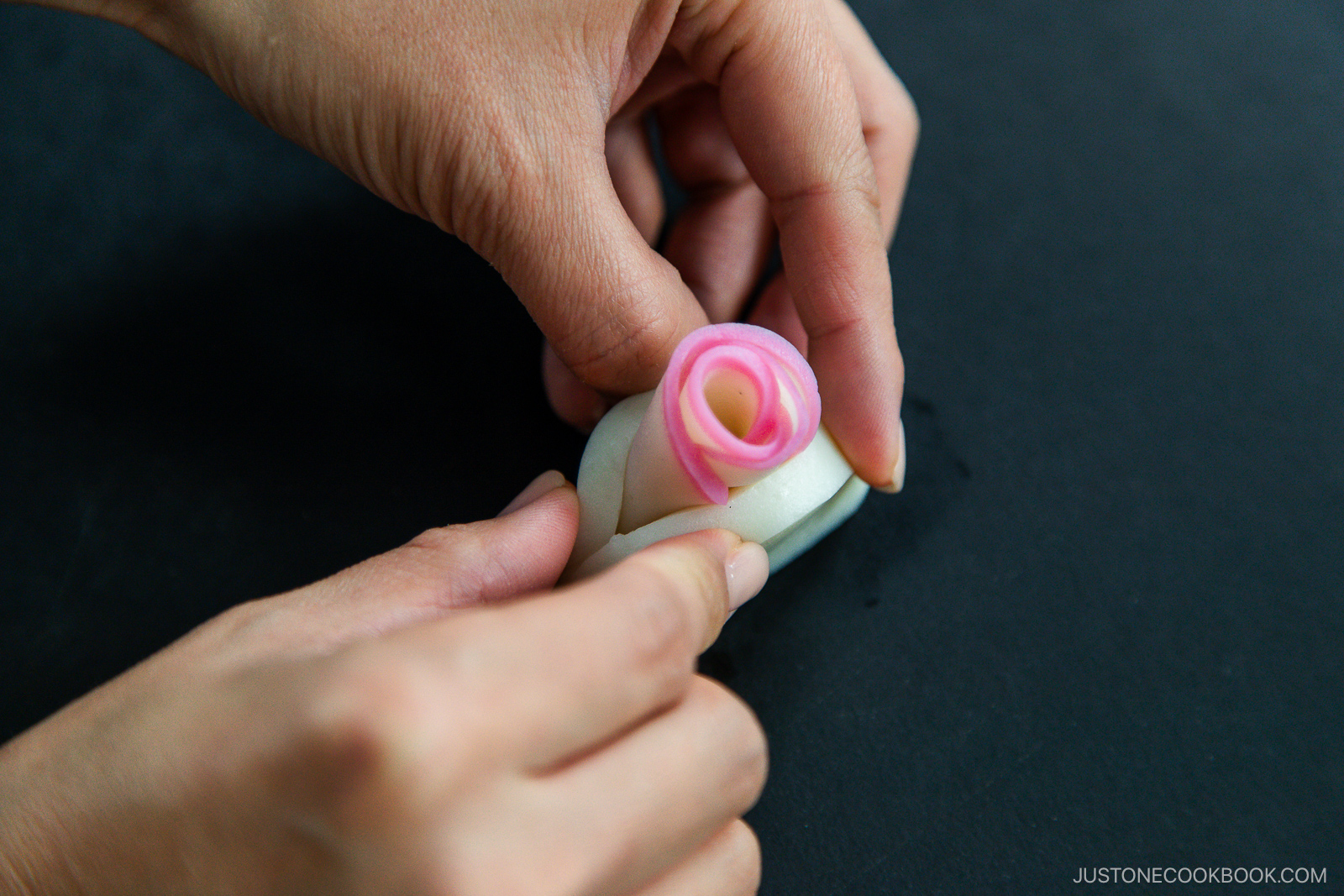 Close-up of hands assembling a delicate edible flower from thinly sliced daikon and pink pickled ginger on a dark surface.
