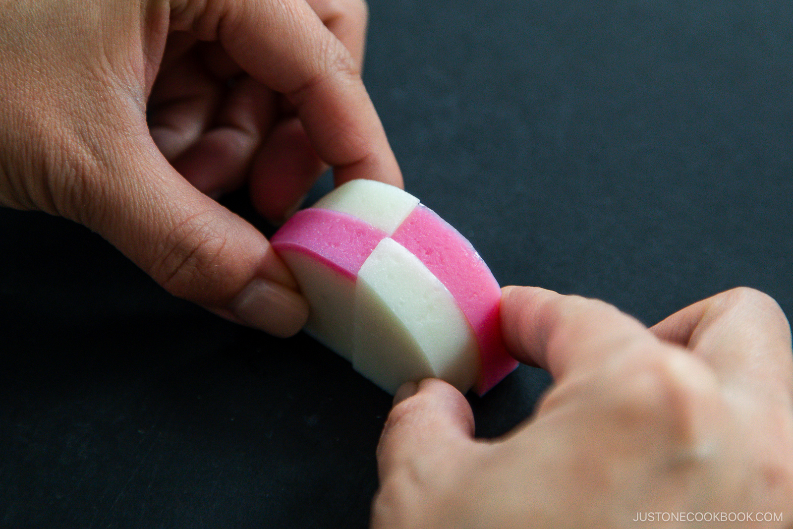 Two hands hold a piece of checkered pink and white Japanese fish cake (kamaboko) against a dark background.