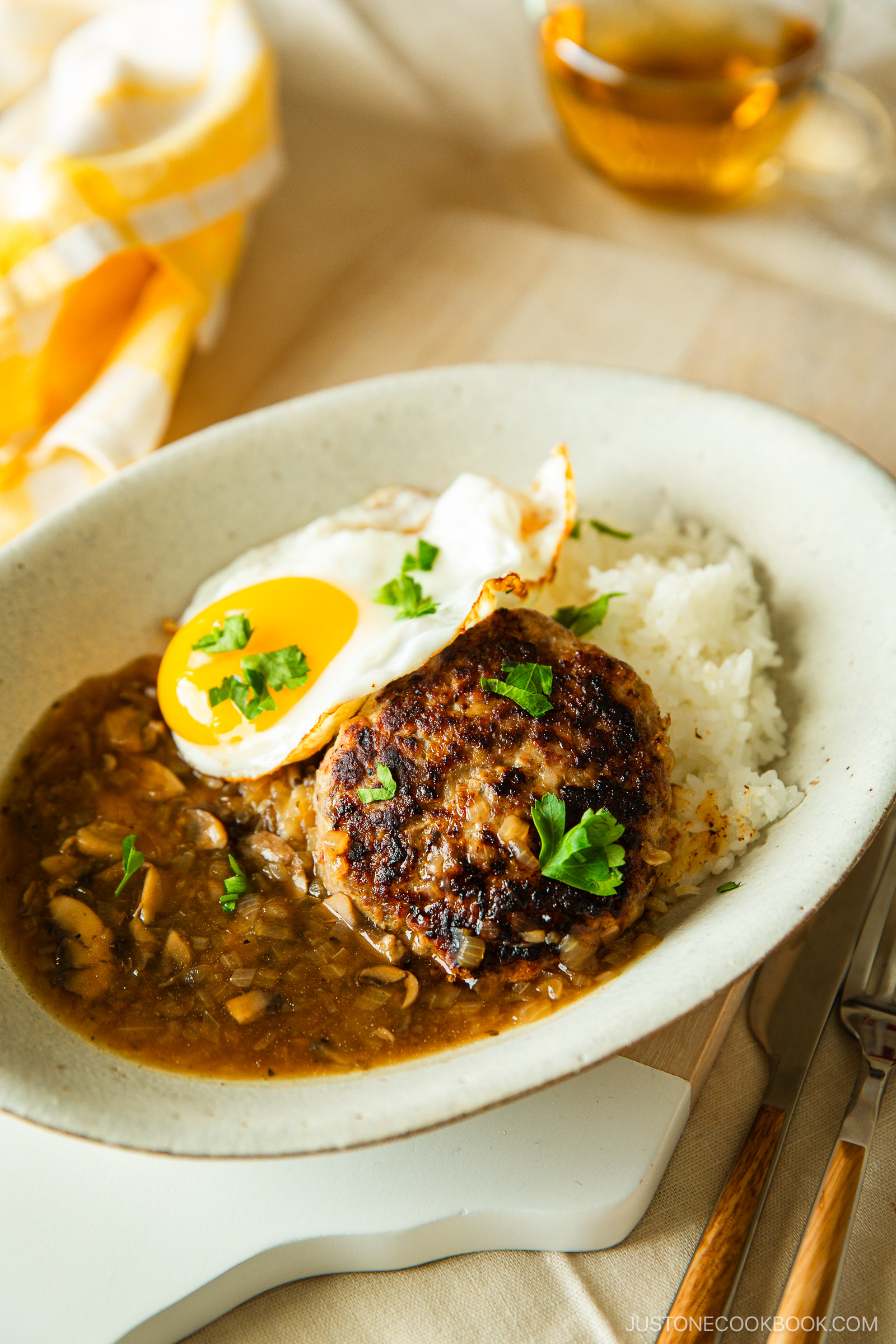 An oval plate with white rice, a browned hamburger patty, a sunny-side-up egg, brown mushroom gravy, and chopped parsley, set on a light surface with a fork, knife, and a cup of tea in the background.